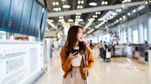 Young Asian Woman In International Airport, Using Mobile Smartphone And Checking Flight At The Flight Information Board