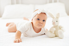Smiling Baby Girl With Blue Eyes Close-up Portrait, Happy Little Baby Of Six Months Lying On Her Tummy With A Stuffed Rabbit In A Bright Bedroom In A White Bodysuit On The Bed And Laughing
