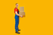 Full-length Portrait Of Smiling Delivery Man In Blue Uniform With Boxes Isolated On Yellow Background. Copy Space, Mock Up.
