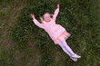 © somemeans - Little smiling girl in a pink dress lies on a green lawn with her arms outstretched and resting. Spring holidays