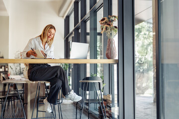 Wall Mural - Smiling business woman working on laptop, making notes in cozy coworkign space interior