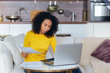 african american woman sitting at home on the couch looking through bills, checking household accoun
