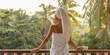 Good morning. Back view of a happy woman with a cup of coffee or tea in a white towel after showering, standing on an open resort balcony against a nature background during a perfect morning in a