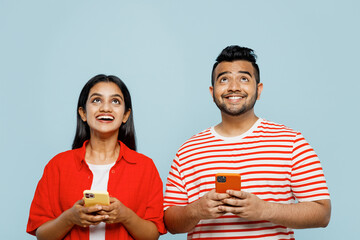 Wall Mural - Young couple two friends family Indian man woman wear red casual clothes t-shirts together hold in hand use mobile cell phone look overhead isolated on pastel plain light blue cyan color background.