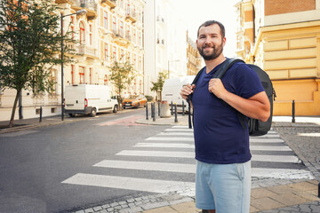 Wall Mural - a handsome bearded man with a backpack in the european center looks at the camera and smiles against the backdrop of beautiful buildings, architecture.Tourism, summer holidays, backpack