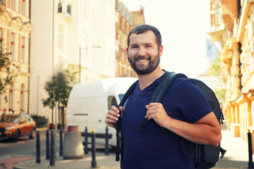 Wall Mural - a handsome bearded man with a backpack in the european center looks at the camera and smiles against the backdrop of beautiful buildings, architecture.Tourism, summer holidays