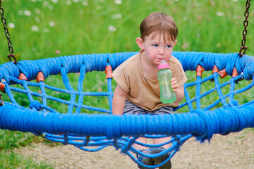 The child swings at the park and drinks water from a bottle with a nipple. Kid aged two years