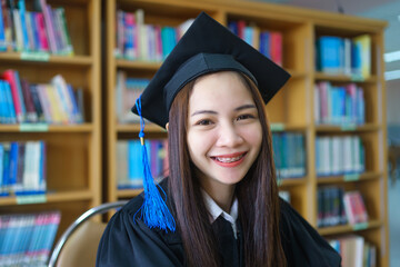 Wall Mural - Portrait of a young happy and excited Asian woman university graduate in academic gown and cap in the library.