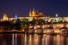 Night Time View Of Charles Bridge Across The Vltava River In The Heart Of Prague With St. Nicholas Church To The Left And Prague Castle And St. Vitus Cathedral Lit Up On The Hilltop In The Background