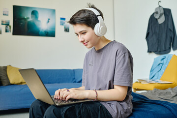 Wall Mural - Brunette female teenager in headphones and casualwear typing on laptop keyboard and looking at screen while sitting on bed and networking