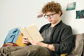 Sticker - Serious teenage guy in eyeglasses and casualwear reading book while sitting on bed against wall woth posters, postcards and photos