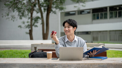 Wall Mural - A happy Asian male college student is doing his homework at a table in a campus park.