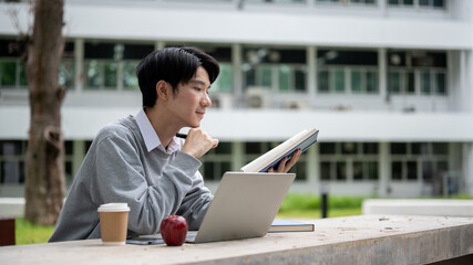 Wall Mural - A smart and focused Asian male university student is reading a book in a campus park.