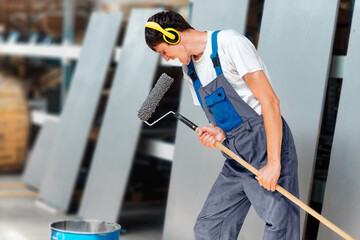 Painter in headphones listens to music and sings into paint roller as if into microphone. Caucasian worker in overalls in good mood relaxes, has fun and sings.