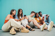 © Jose Calsina - Multiracial smiling teenagers having fun sharing on the social media with their smart phones sitting in row on the floor, blue background . Young multiethnic students enjoying and laughing with funny