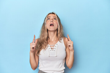 Wall Mural - Blonde young caucasian woman in blue studio pointing upside with opened mouth.