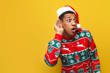 african american guy in new year clothes and santa hat eavesdrops and wonders on yellow isolated background