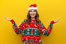 Girl In Christmas Sweater And Santa Claus Hat Spreads Her Arms To The Sides On Yellow Isolated Background