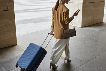 Rear view of young modern businesswoman pulling suitcase and texting in mobile phone while moving along columns of architecture to airport