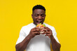 © Богдан Маліцький - african american man in white t-shirt bites big burger on yellow isolated background, young guy eats fast food
