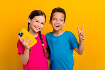 Sticker - Photo of excited pretty preteen schoolkids dressed t-shirts texting modern gadget showing v-sign isolated yellow color background