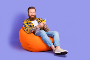 Wall Mural - Photo of smiling handsome man wear checkered stylish clothes sitting pouf use modern phone empty space isolated on purple color background