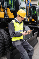 Aufkleber - Engineer in a helmet with a digital tablet stands next to construction excavators.