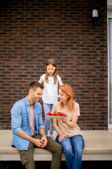 Wall Mural - Family with a mother, father and daughter sitting outside on steps of a front porch of a brick house and eating strawberries