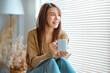 © oatawa - Joyful young asian female enjoying a cup of coffee while sitting on bench at home, Cosy scene, Smiling pretty woman drinking hot tea in autumn winter.
