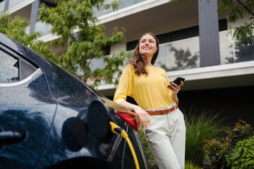 Close up of beautiful woman using smartrphone while charging her electric car on the street.