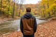 © Florian - A man standing in front of a river in a forest, gray and brown hoodie. The river is shallow and rocky, with a few small rapids, contemplative, peaceful