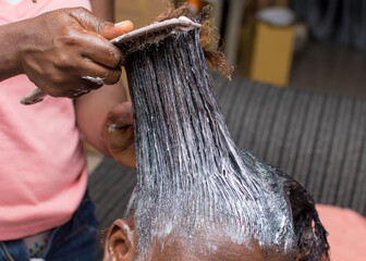 Hands of an African stylist, holding and applying relaxer cream with a comb to the natural long hair of a woman or female customer at a beauty salon in Nigeria