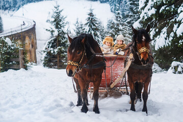 Two children ride on a retro sleigh with horses in winter time. Mountain snowy landscape background. Tours, horse riding.