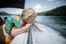 Young Child Leaning Over Boat Railing To Touch Wake On Lake