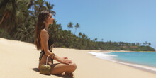 Side View Of Bikini Woman In Black Swimsuit Relaxing And Sunbathing And Meditating On Tropical Beach Near The Ocean. Attractive Brunette Girl In Sitting With Coconut On A Perfect Sandy Coast With Palm