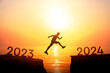 Silhouette of young active man jumping on cliff from number 2023 to 2024 years over the precipice and ocean with sunset or sunrise sky background. New year's concept. End of 2023 Happy New Year 2024.