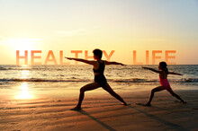 Silhouette Of Mom And Daughter Practicing Yoga Warrior II Pose On Tropical Beach With Sunset Sky Background, Watching The Sunset, Standing As A Part Of The Wording Concept For Healthy Life.
