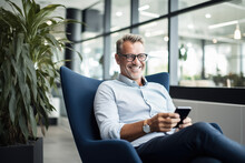 Smiling Mid Aged Business Man Sitting On Chair In Modern Office Space Using Cell Phone Services And Solutions. Mature Businessman Professional Holding Mobile Working On Smartphone Technology