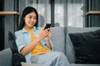 © photobyphotoboy - Smiling young asian woman using mobile phone checking social media,app playing game, shopping online, ordering delivery relax  while sitting on a couch at home with laptop computer