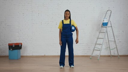 Full length shot of an exhausted dark-skinned young female construction worker standing in the room under renovation, with her hardhat under her arm.