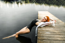 Young Sexy Woman With Long Red Hair In A White Shirt Posing On A Wooden Pier. Young Beautiful Redhead Woman With Long Wet Hair Relaxing In The Water Of A Lake.