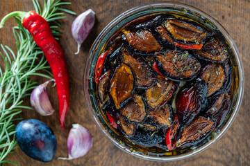 Wall Mural - Sun-dried plums with garlic, green rosemary, red chili pepper, olive oil and spices in a glass jar on a wooden table. Rustic style, top view, close up