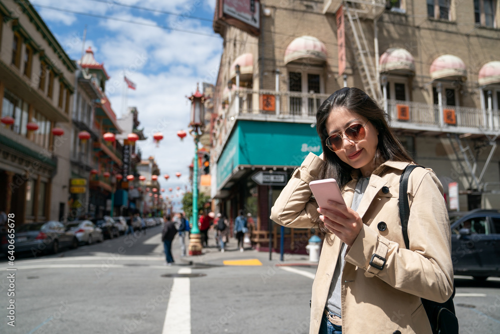 smiling asian Japanese traveler wearing sunglasses and reading guide on cellphone at street corner while visiting Chinatown neighborhood in san Francisco California usa on a sunny day