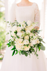 Wall Mural - A bride in a classic long-sleeved lace wedding dress is holding a traditional bouquet with white roses and white flowers and green eucalyptus leaves 