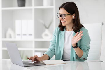 Wall Mural - Glad millennial european businesswoman in glasses uses computer to chat, waving hand