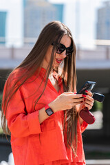 Poster - Summer portrait of a girl with long blond hair. A girl in a orange shirt and sunglasses look at the smartphone screen.