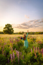 Beautiful Woman In A Straw Hat And Bright Dress With A Bouquet Of Purple Lupins Flowers. Smiling Woman Enjoys The Weather And Scenery While Walking In The Countryside.