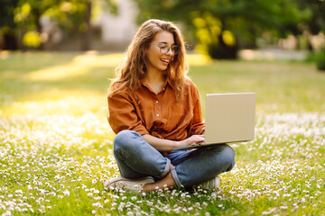 Wall Mural - Portrait of a young woman sitting on the grass in the park working, studying on a laptop. Happy woman enjoys the sunny weather in the meadow. Freelancing concept, online learning.