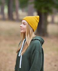 Wall Mural - Attractive blue eyed blonde woman walk on the park. Girl wear green hoodie and yellow hat. Portrait of a joyful young woman enjoying in autumn park.