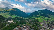 Forwards Fly Over Beautiful Alpine Landscape With Town, Hills And Deep Valleys. Aerial Hyperlapse Shot With Clouds Rolling In The Sky. Gstaad, Switzerland
